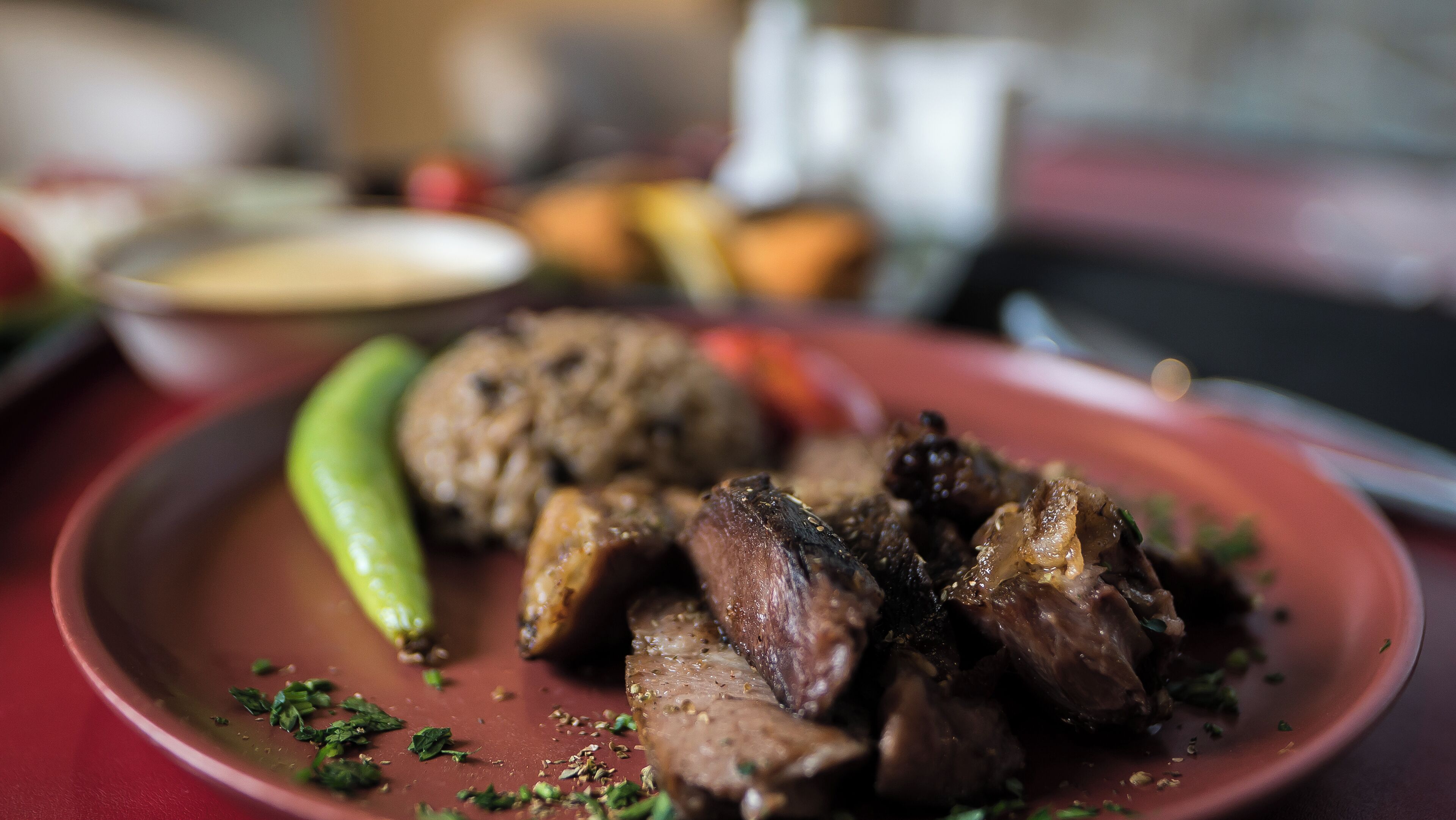 Un plato con carne en rodajas y un chile verde, adornado con hierbas, sobre un mantel rojo.