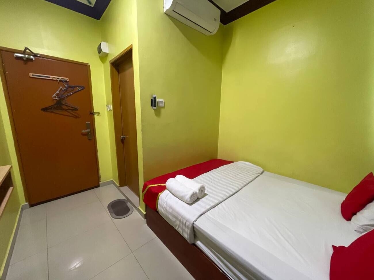 Una habitación pequeña con cama individual, cubierta con un edredón rojo y blanco, una puerta de madera con perchero para toallas y una unidad de aire acondicionado instalada en el techo.