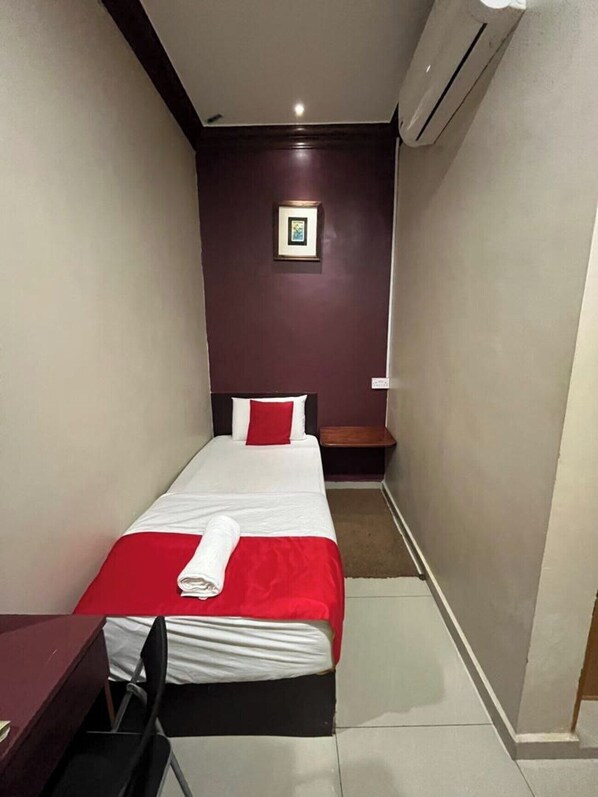A small, narrow hotel room with a single bed, a desk, and a chair.