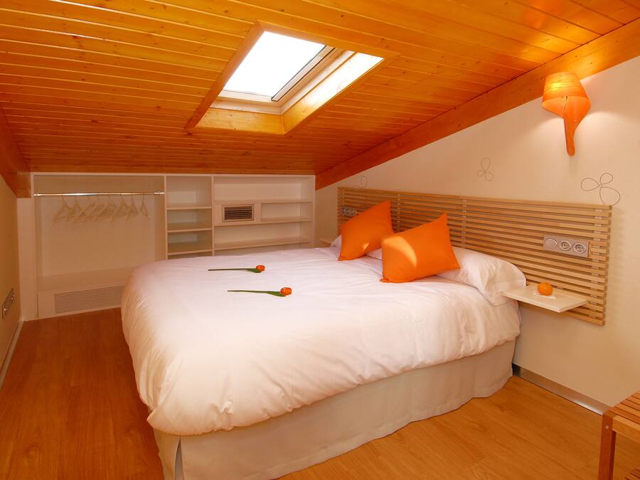 A bedroom with a skylight, wooden ceiling, white bedding, orange pillows, and a bedside table with a fruit.