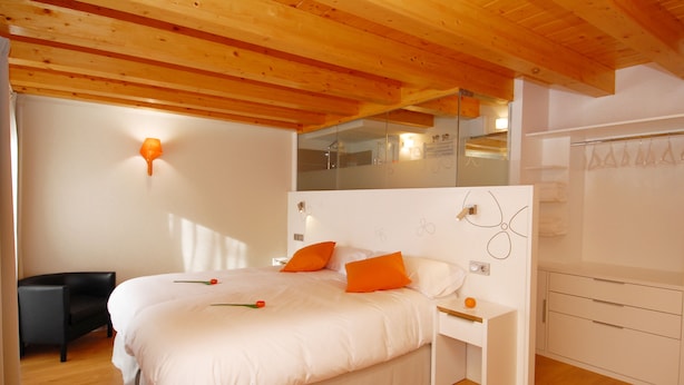 A bedroom with a wooden ceiling, white walls, a bed with orange pillows, a nightstand, and a mirror. - Hostal riMboMbin (Burgos)