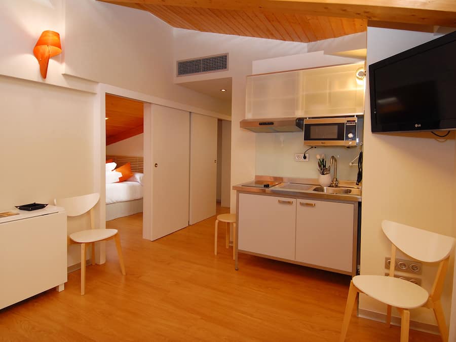 Una cocina compacta con microondas, estufa y pileta, un pequeño comedor con dos sillas, y un televisor montado en la pared.