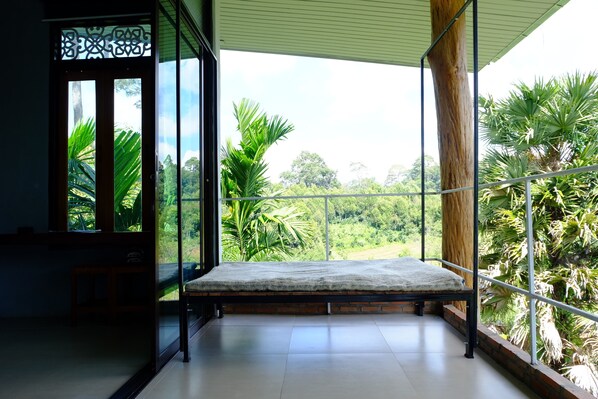 A balcony with a bed, a view of lush greenery, and a wooden column.