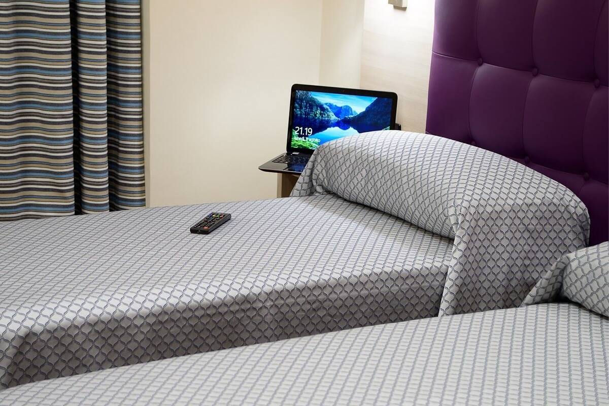 Une chambre d’hôtel avec un lit, un ordinateur portable posé sur une table de chevet et une tête de lit violette.