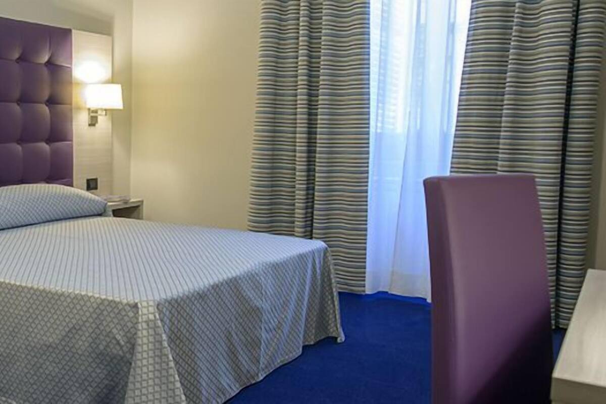 Une chambre d’hôtel avec un lit, une table de chevet, une lampe, une chaise et une fenêtre avec des rideaux.
