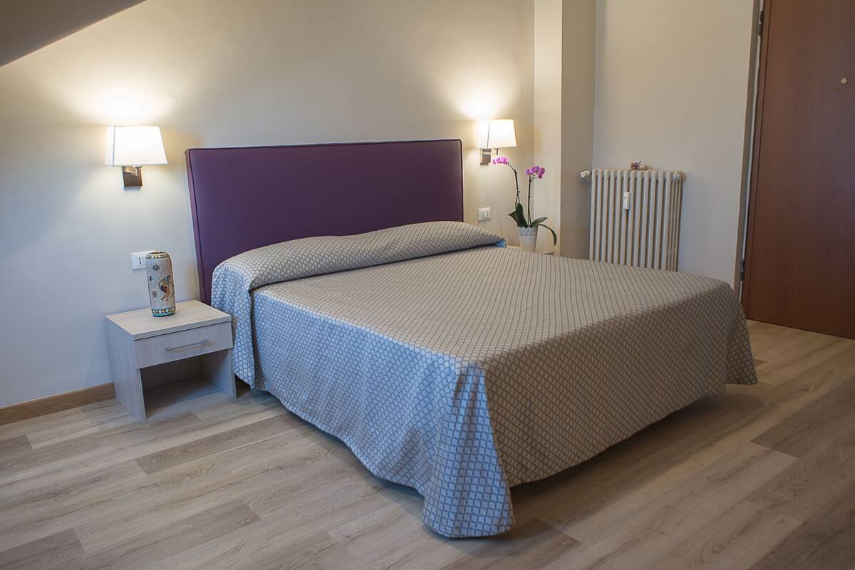 Un lit bien fait, avec une tête de lit violette, une table de chevet sur laquelle se trouve une lampe, et un radiateur fixé au mur.