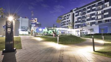 A well-lit urban area with a modern building, a grassy hill, and a construction site in the background.