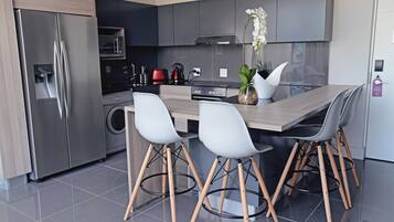 Una cocina moderna con un refrigerador de acero inoxidable, un microondas, una estufa y un lavamanos. Hay dos sillas blancas con patas de madera y una mesa de madera.