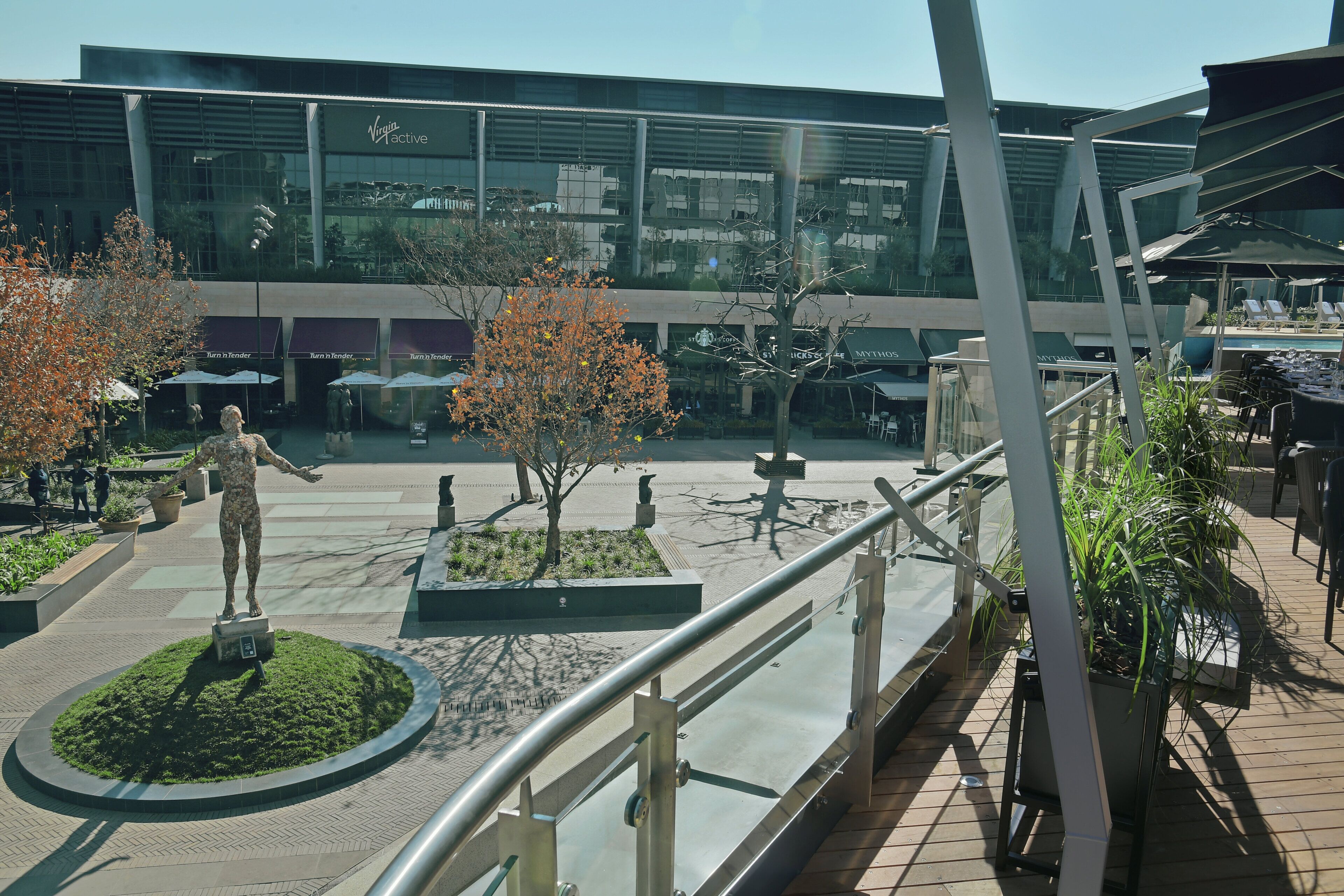 A modern building with a glass facade, outdoor seating areas, and a statue in the center.