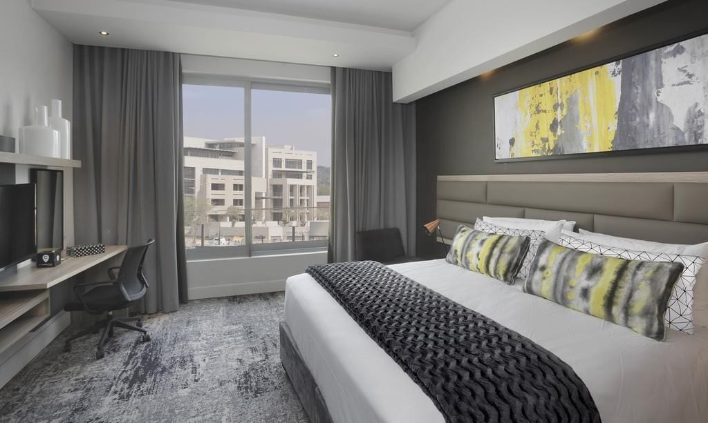 A hotel room with a large bed, a desk, a chair, and a view of buildings through the window.