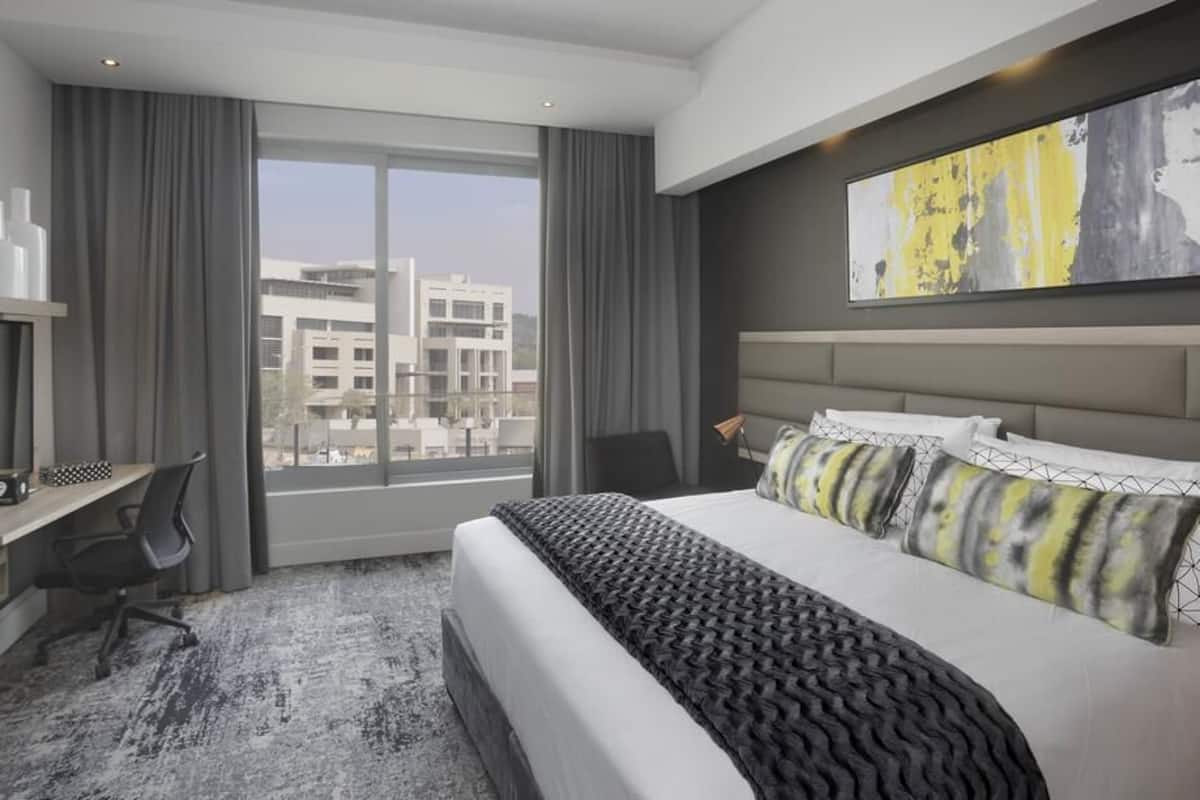 A modern hotel room with a large bed, a desk, and a view of buildings through the window.