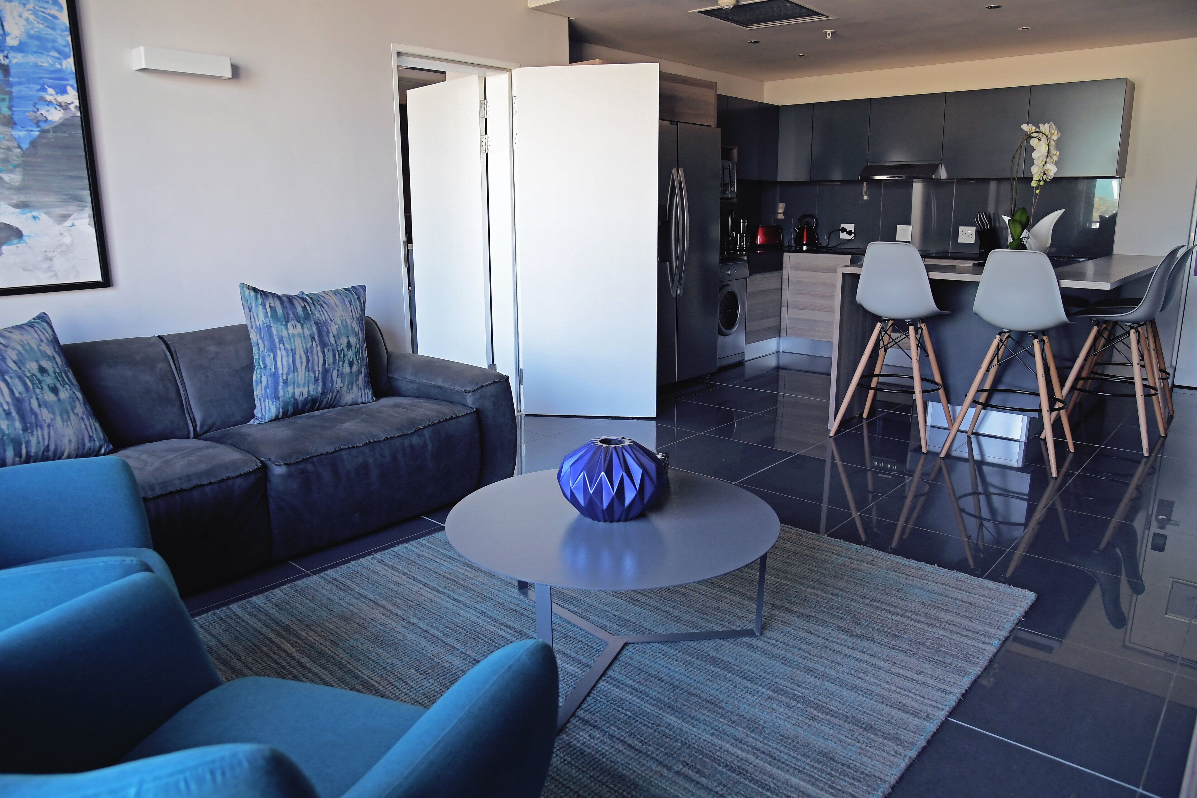 Een moderne woonkamer met een bank, salontafel en een keukenhoek met een koelkast, spoelbak en barstoelen.