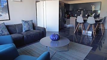 Een moderne woonkamer met een bank, salontafel en een keukenhoek met een koelkast, spoelbak en barstoelen.