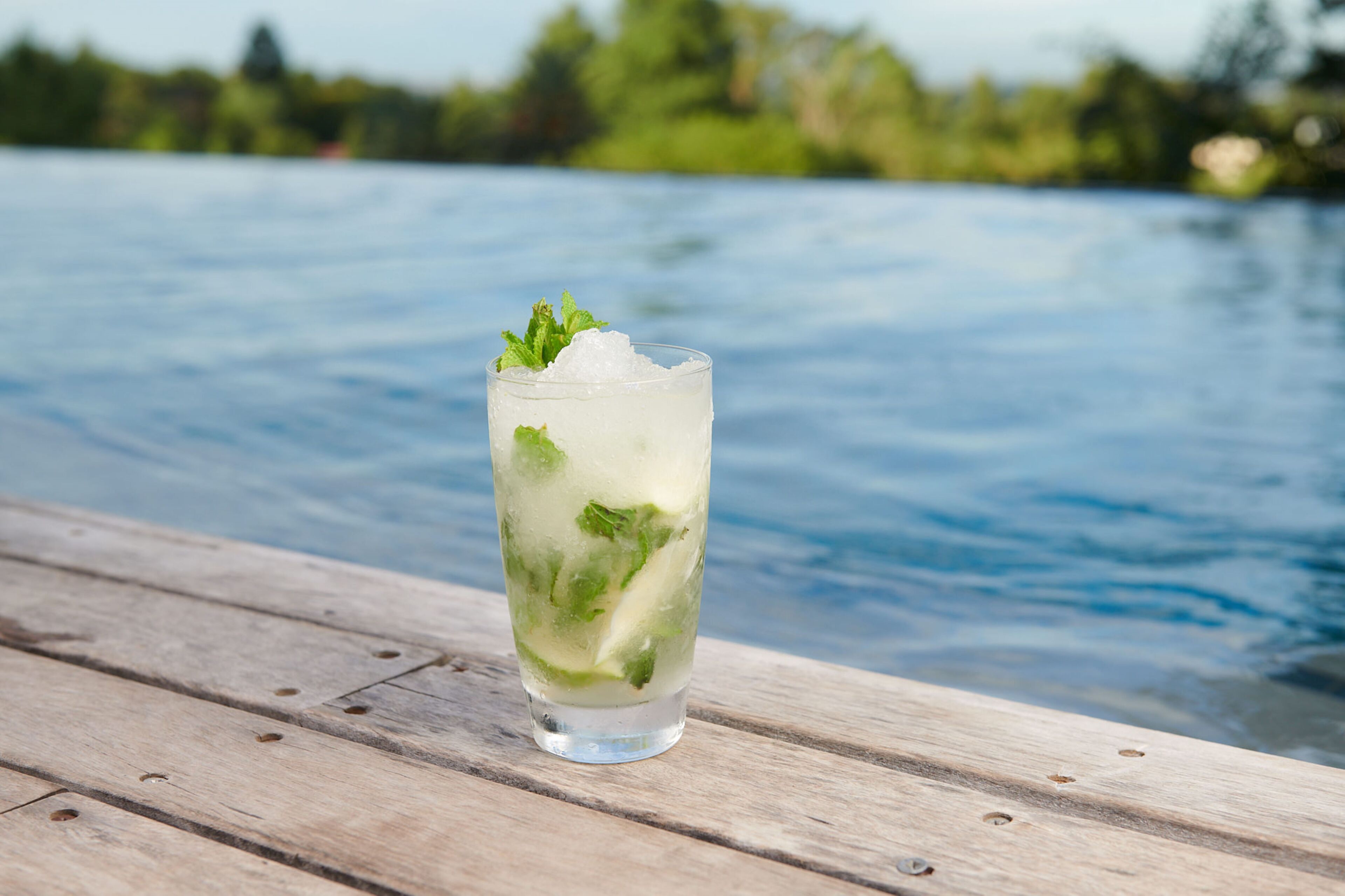 A glass of iced drink with mint on a wooden deck by a body of water.