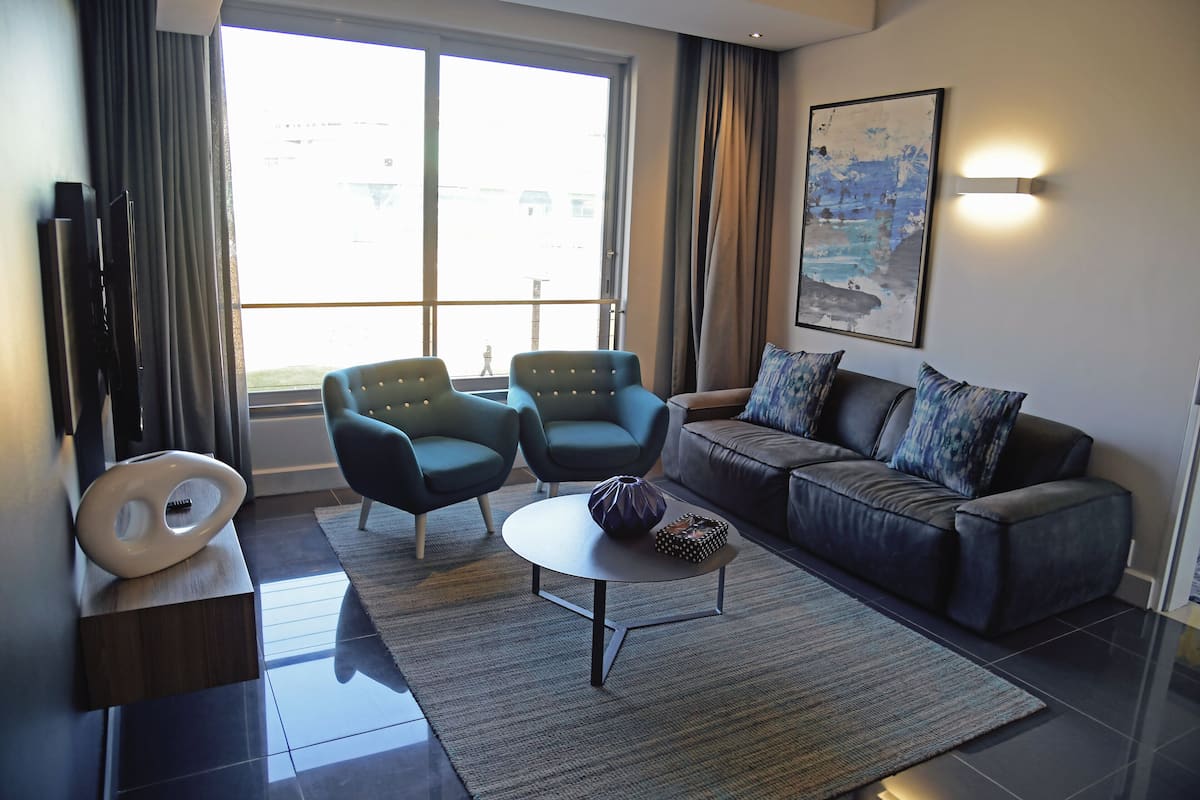 A modern living room with a sofa, two armchairs, a coffee table, a TV, and a large window with curtains.