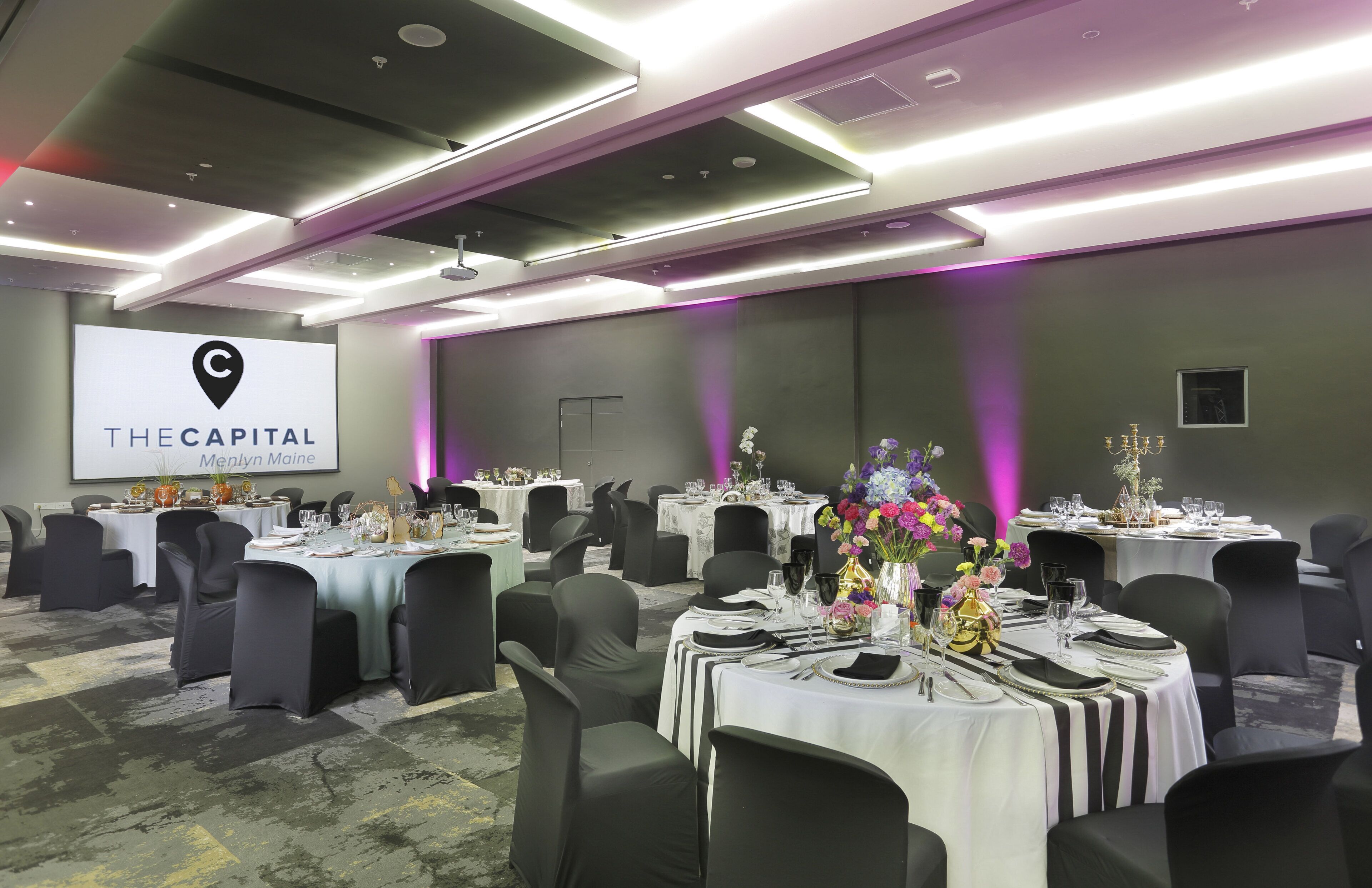 A conference room at The Capital Hotel with tables set for a meeting, black chairs, and a projector screen displaying the hotel name.