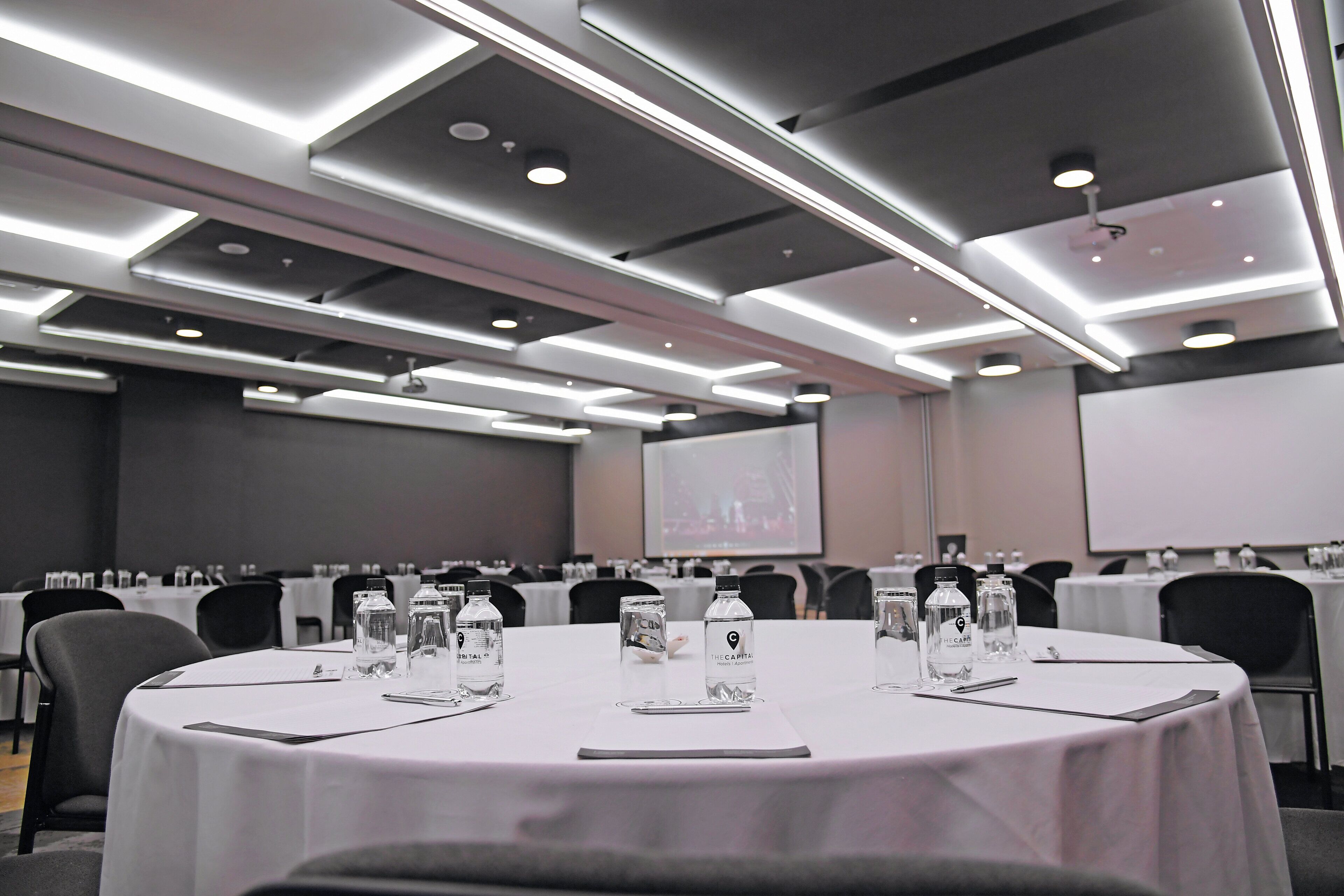 Un salón de conferencias con mesas redondas, botellas de agua y una pantalla de proyector.