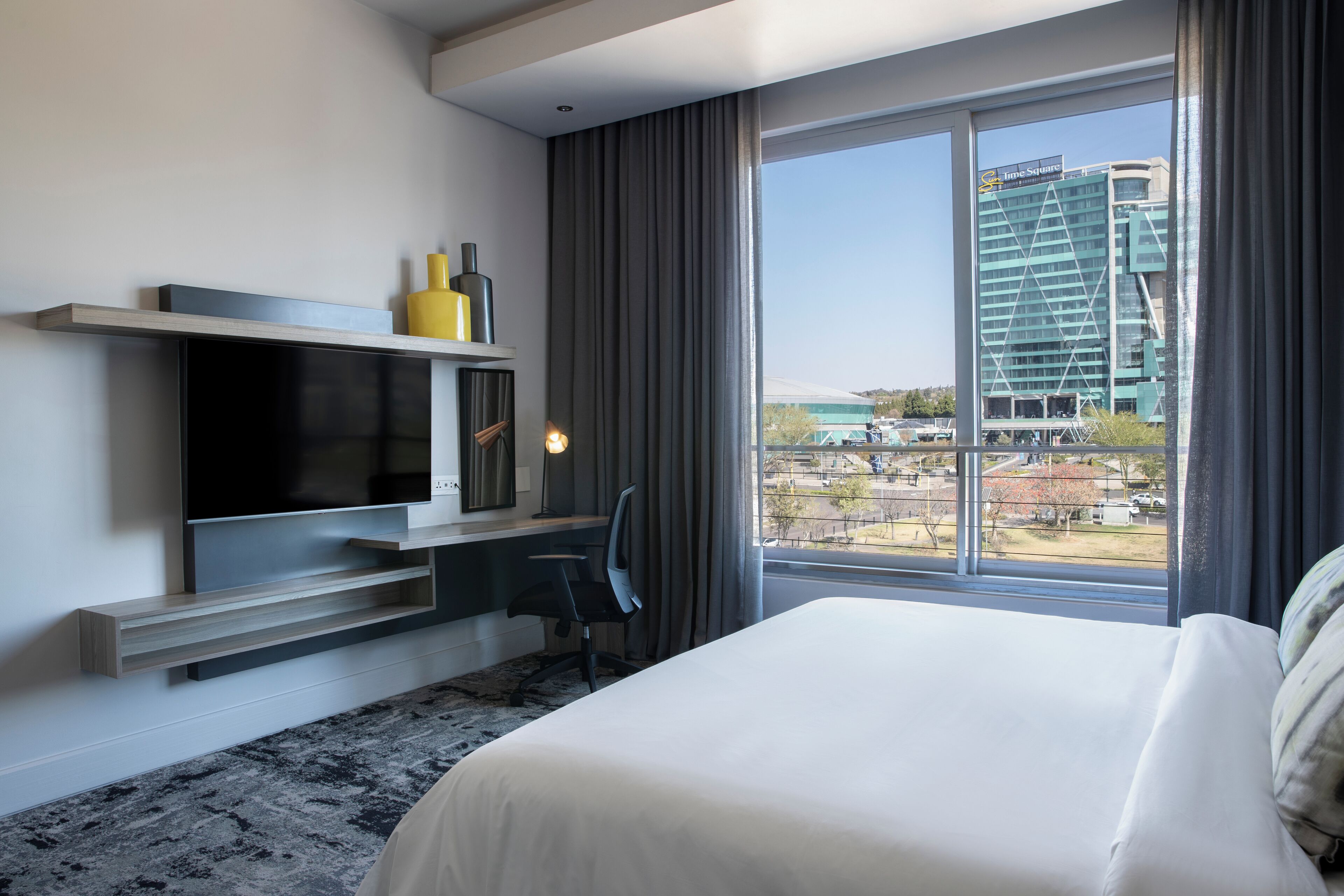 A modern hotel room with a large bed, a flat-screen TV mounted on the wall, a desk with a chair, and a large window offering a view of the cityscape.