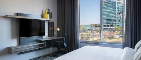 Une chambre d’hôtel moderne dotée d’un grand lit, d’un téléviseur à écran plat fixé au mur, d’un bureau avec une chaise et d’une grande fenêtre donnant sur le paysage urbain.