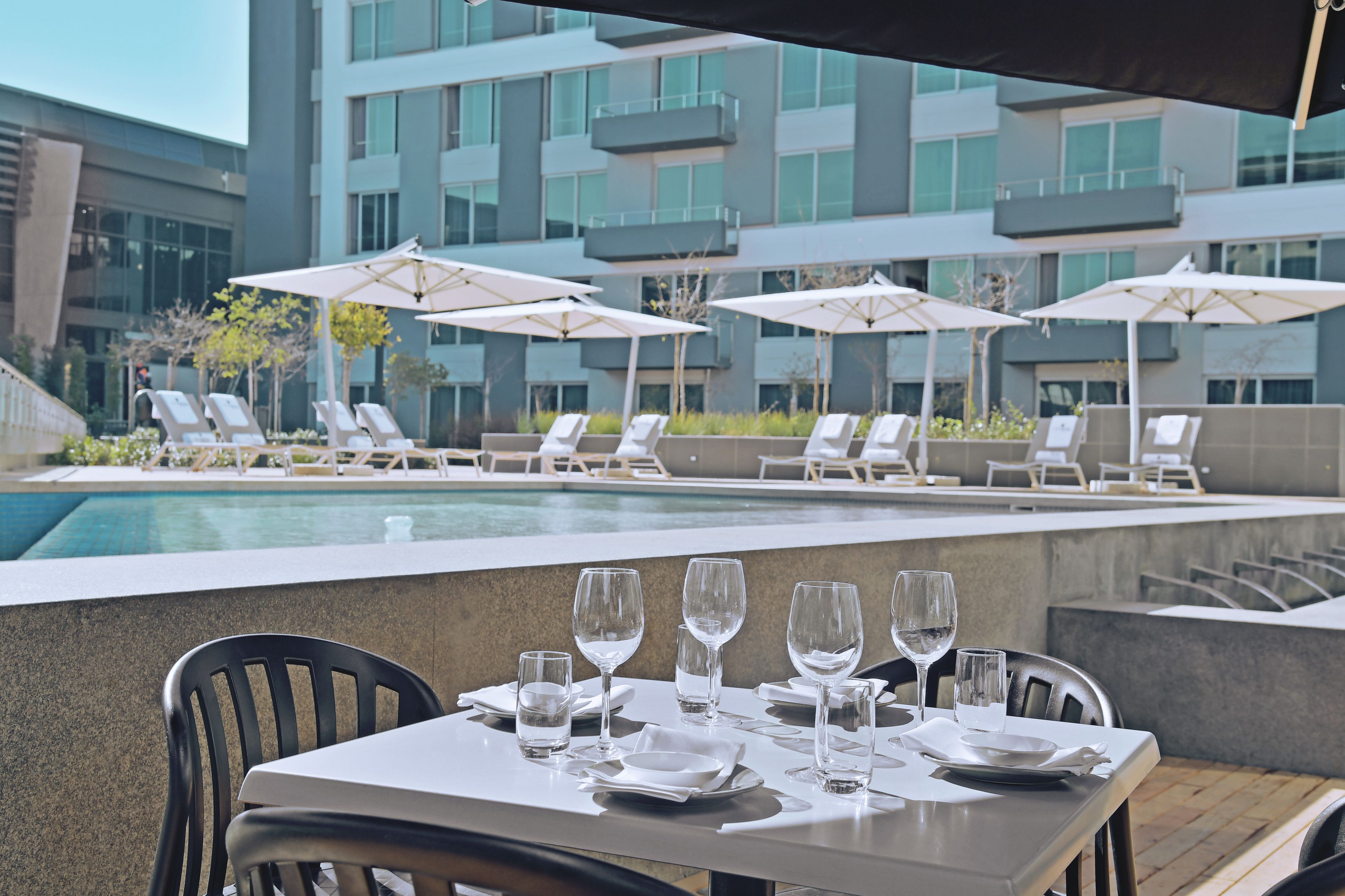 A dining area with tables set for a meal, overlooking a swimming pool and a modern building with balconies.