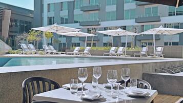 A dining area with tables set for a meal, overlooking a swimming pool and a modern building with balconies.