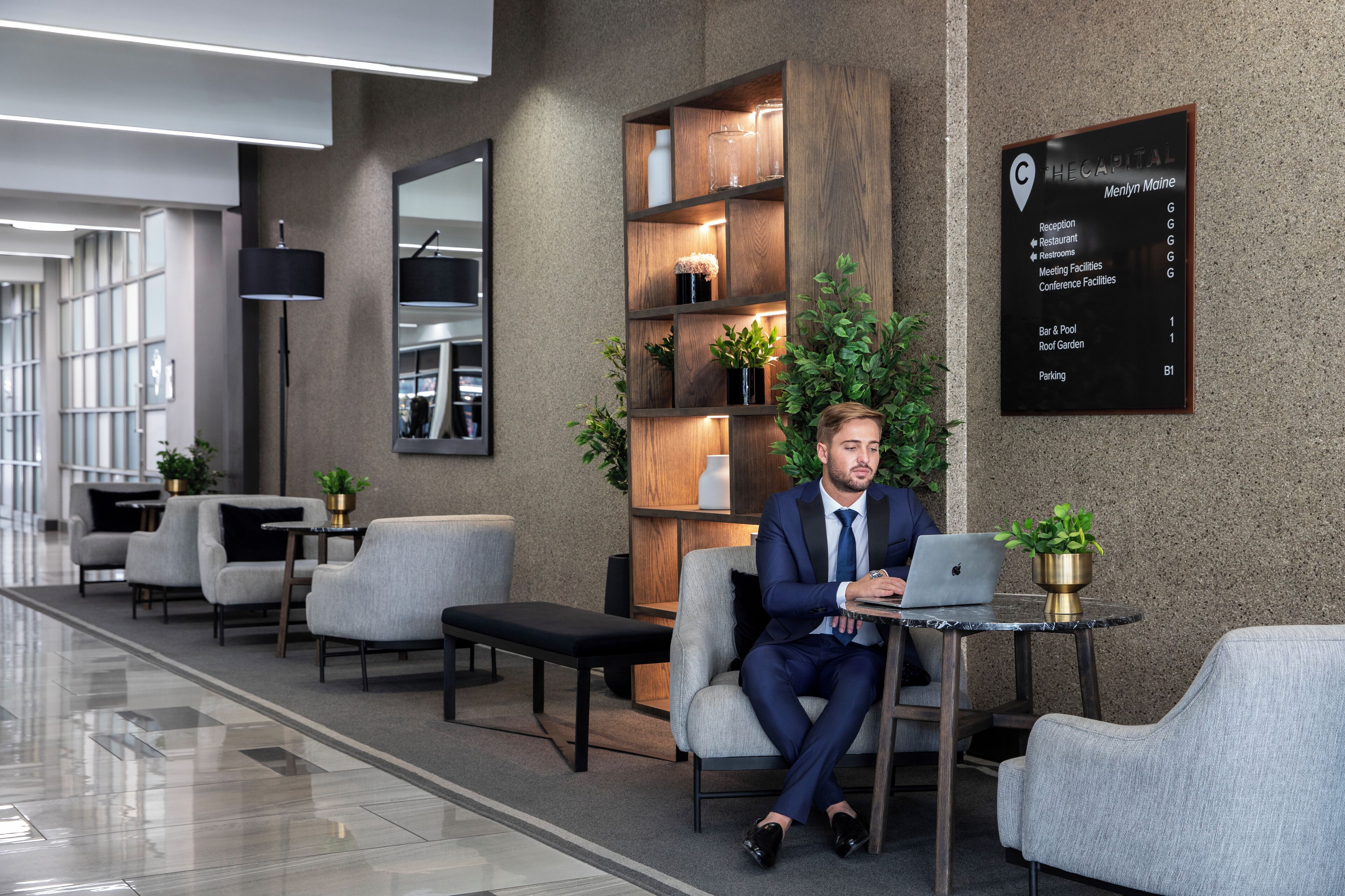 Un hombre con traje azul sentado a una mesa con una computadora portátil en un moderno vestíbulo de hotel, que incluye un mostrador de recepción, una zona de estar y un letrero con indicaciones.