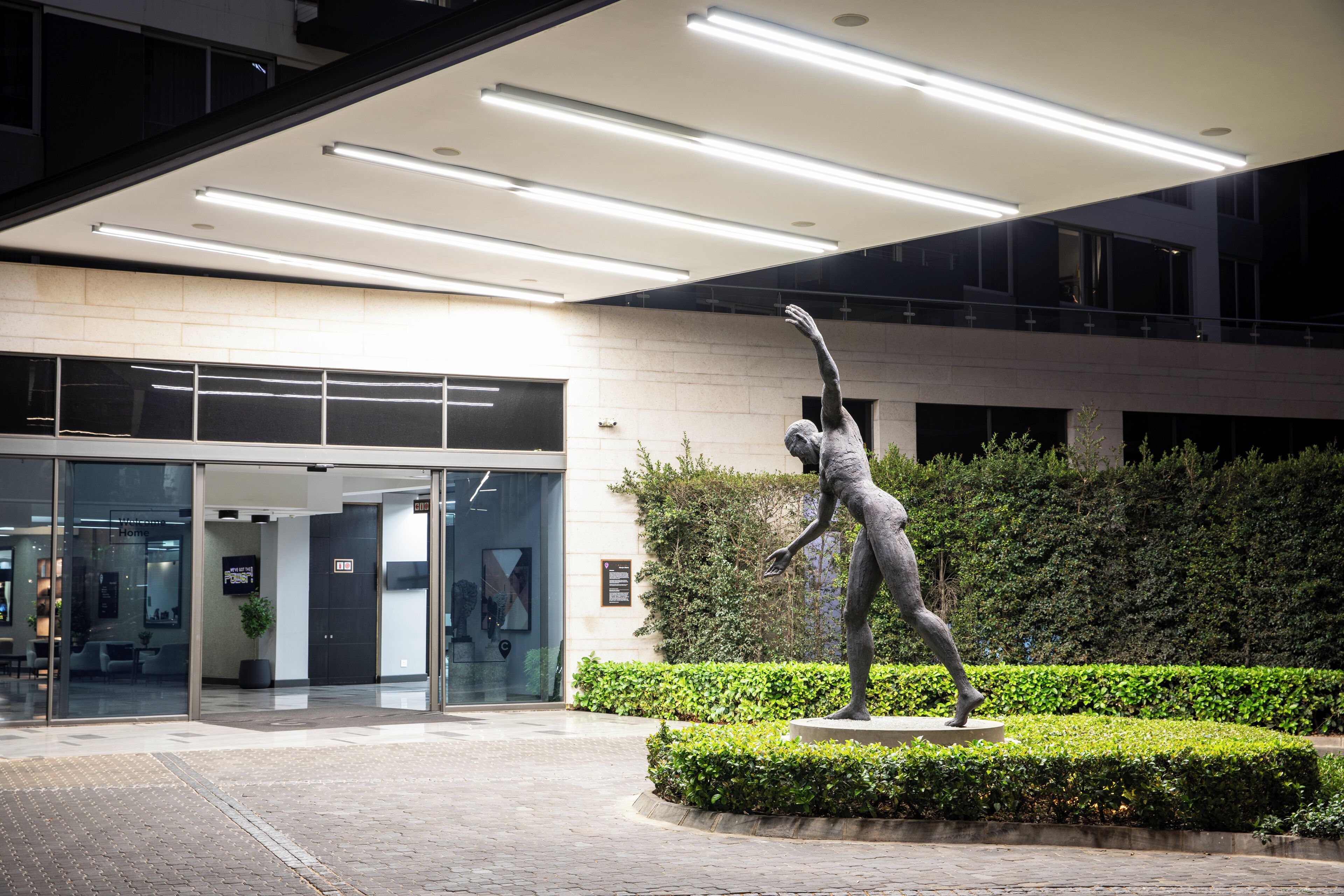 A modern building entrance with a statue of a figure in motion, surrounded by well-maintained greenery.