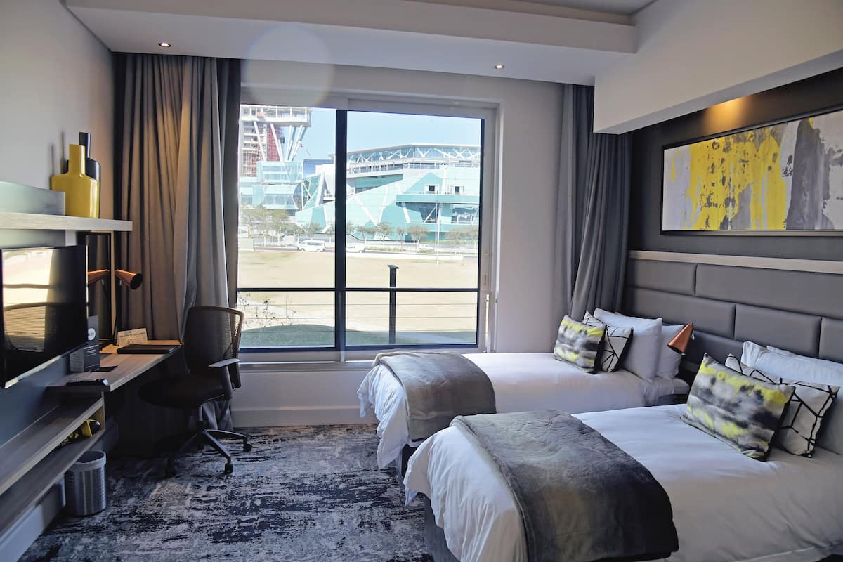 A hotel room with two beds, a desk with a chair, a television, and a large window offering a view of an industrial area.