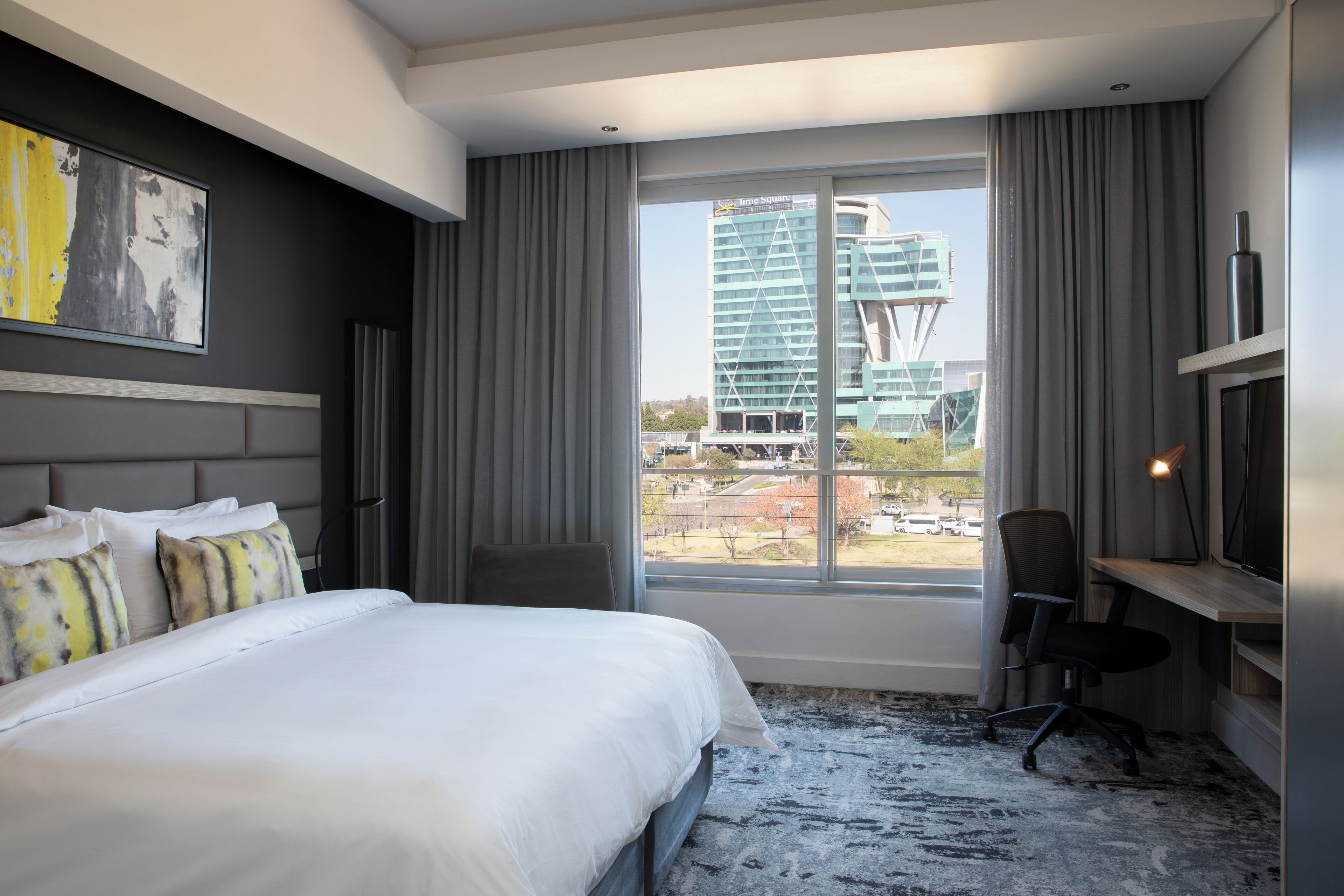 A modern hotel room with a large bed, a desk, and a view of a cityscape.