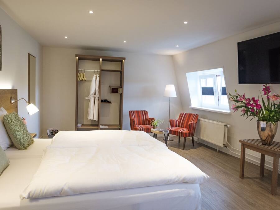A hotel room with a large bed, a seating area with two chairs, a TV mounted on the wall, and a window with flowers on the table.