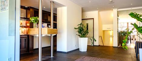 A modern hotel lobby with a reception desk, a large potted plant, and a visible door leading to the bathroom.