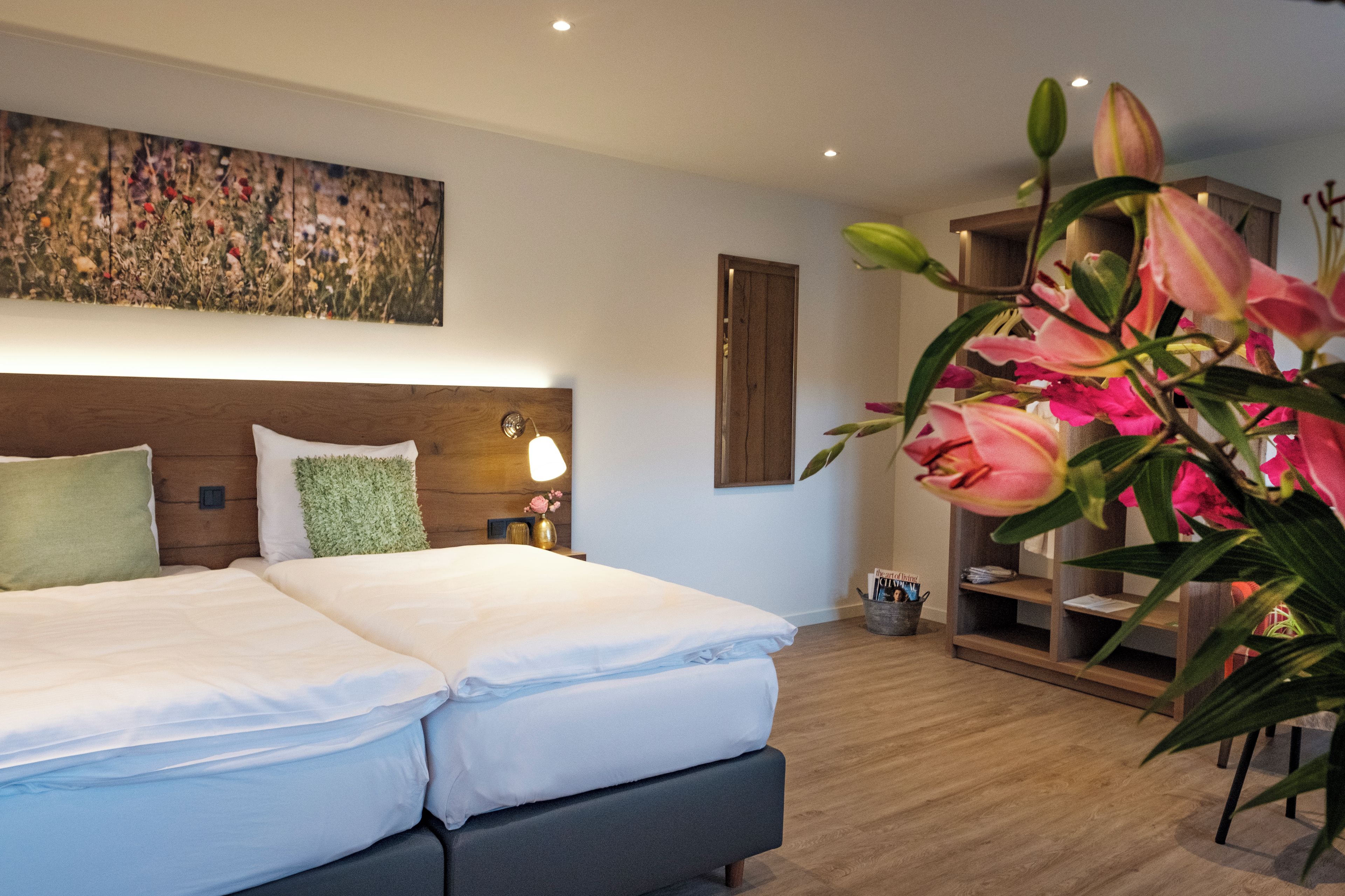 Habitación de hotel con cama, lámparas de noche, mesita auxiliar, estante en la pared y un cuadro grande con motivos florales.