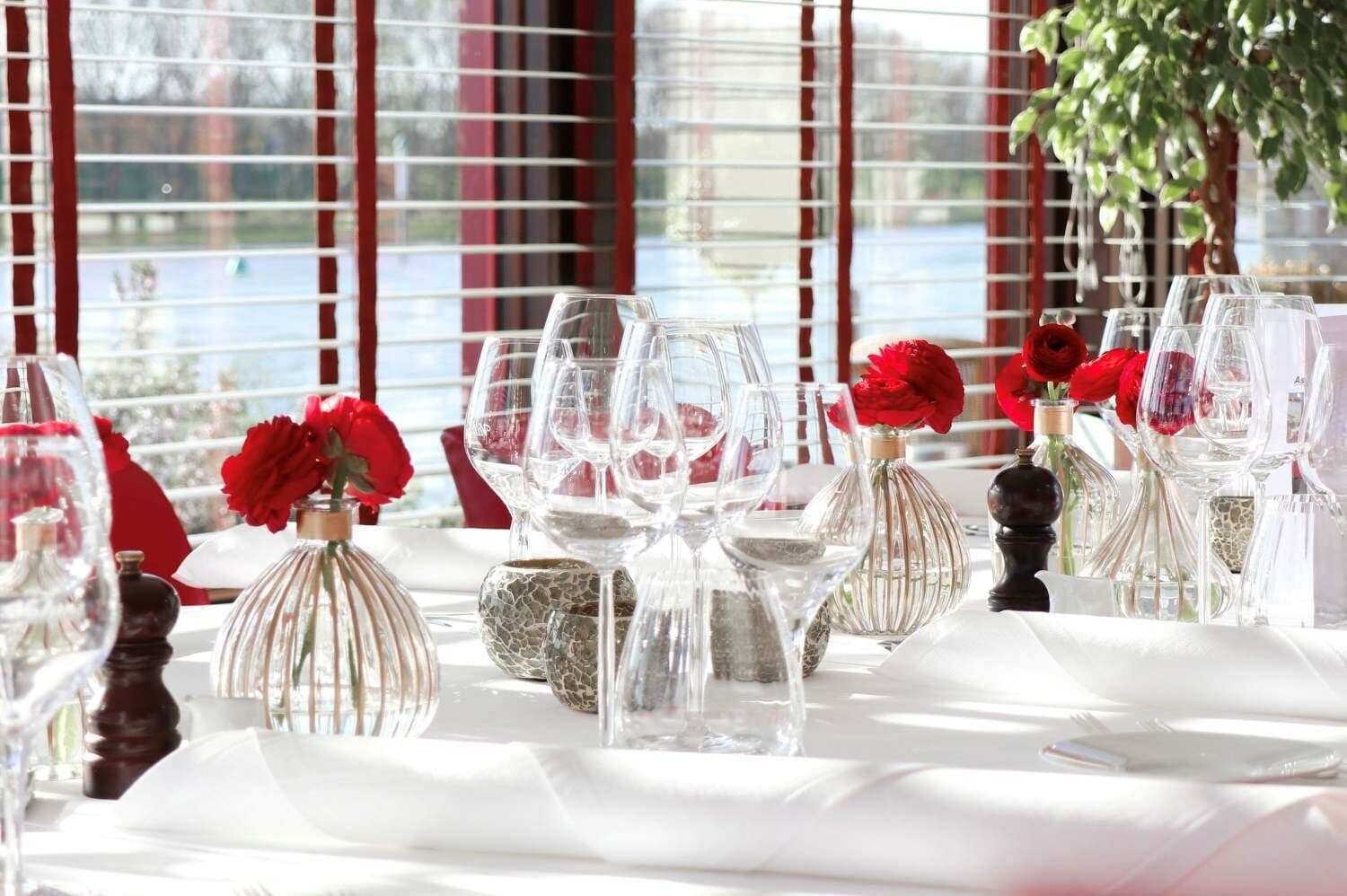 A table set for dining with clear glassware and red roses in vases, set against a window with blinds.