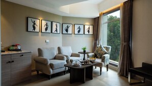 A modern living room with a sofa, armchairs, a coffee table, and a dining table. There are framed pictures on the wall, a window with curtains, and a view of greenery outside. - Fortune Park Sishmo, Bhubaneswar - Member ITC Hotels' Group (Bhubaneshwar)