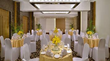 Un salón de banquetes con mesas redondas, sillas blancas, manteles dorados y flores amarillas.