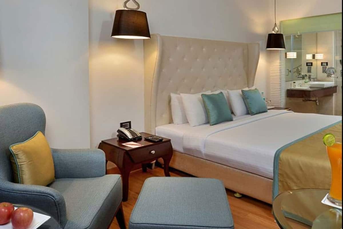 Ett hotellrum med en säng, ett skrivbord med en telefon, en stol, en pall och ett glasbord med ett glas på.