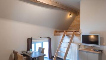 A compact room with a loft bed, a desk with a TV, a dining area, and a window with curtains.