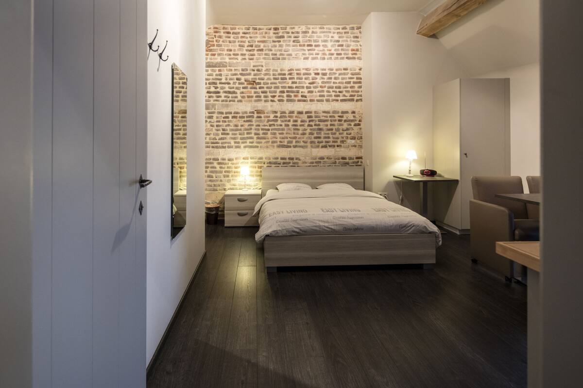 Un dormitorio moderno con cabecera de ladrillo, una cama bien hecha, una mesita de noche con lámpara y una silla.