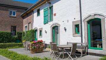 Un edificio blanco con contraventanas verdes y fachada de ladrillo. Hay una mesa y sillas de madera afuera, junto a una jardinera con flores.