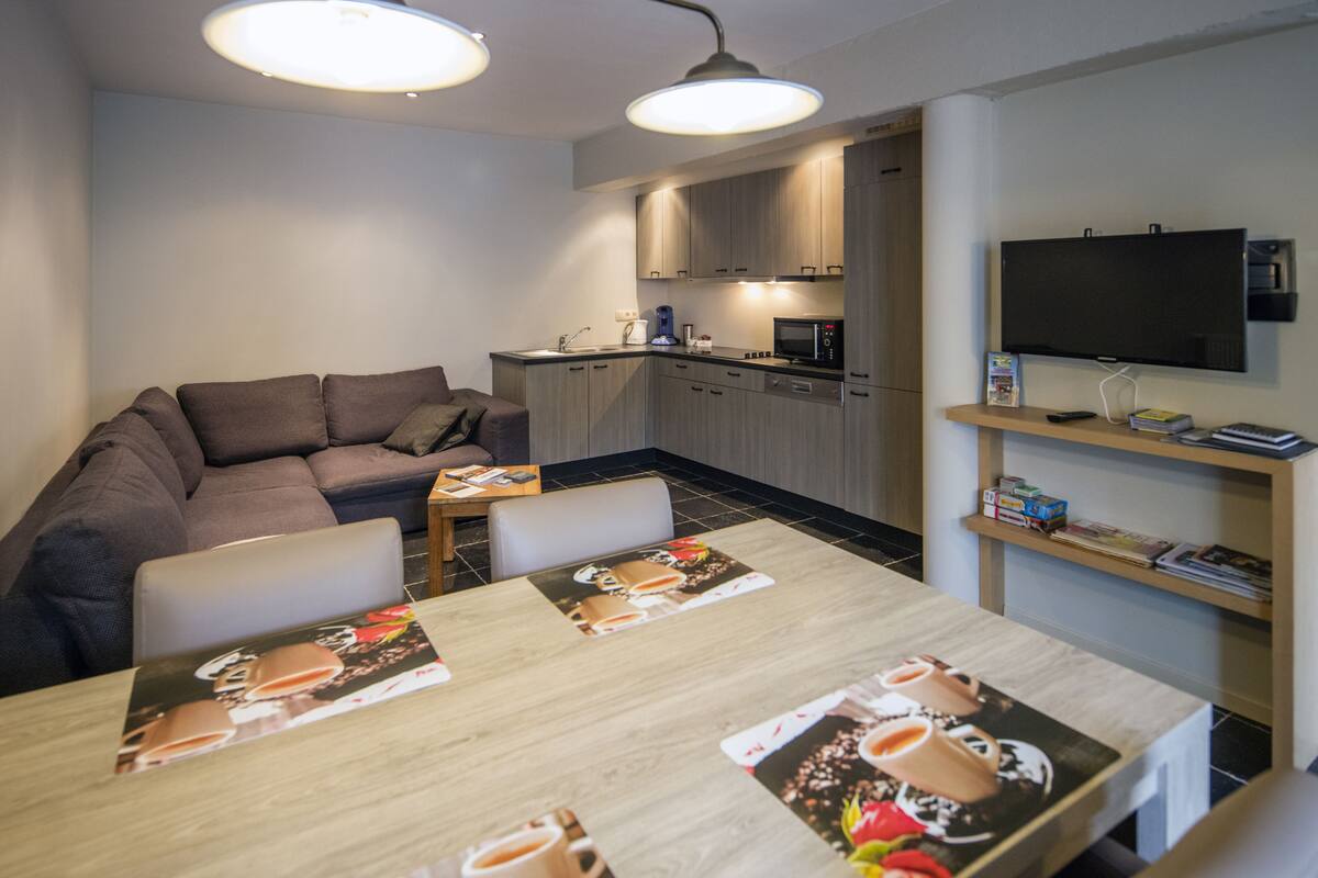 Una sala de estar moderna con un sofá, una mesa de comedor con individuales, una zona de cocina y un televisor montado en la pared.