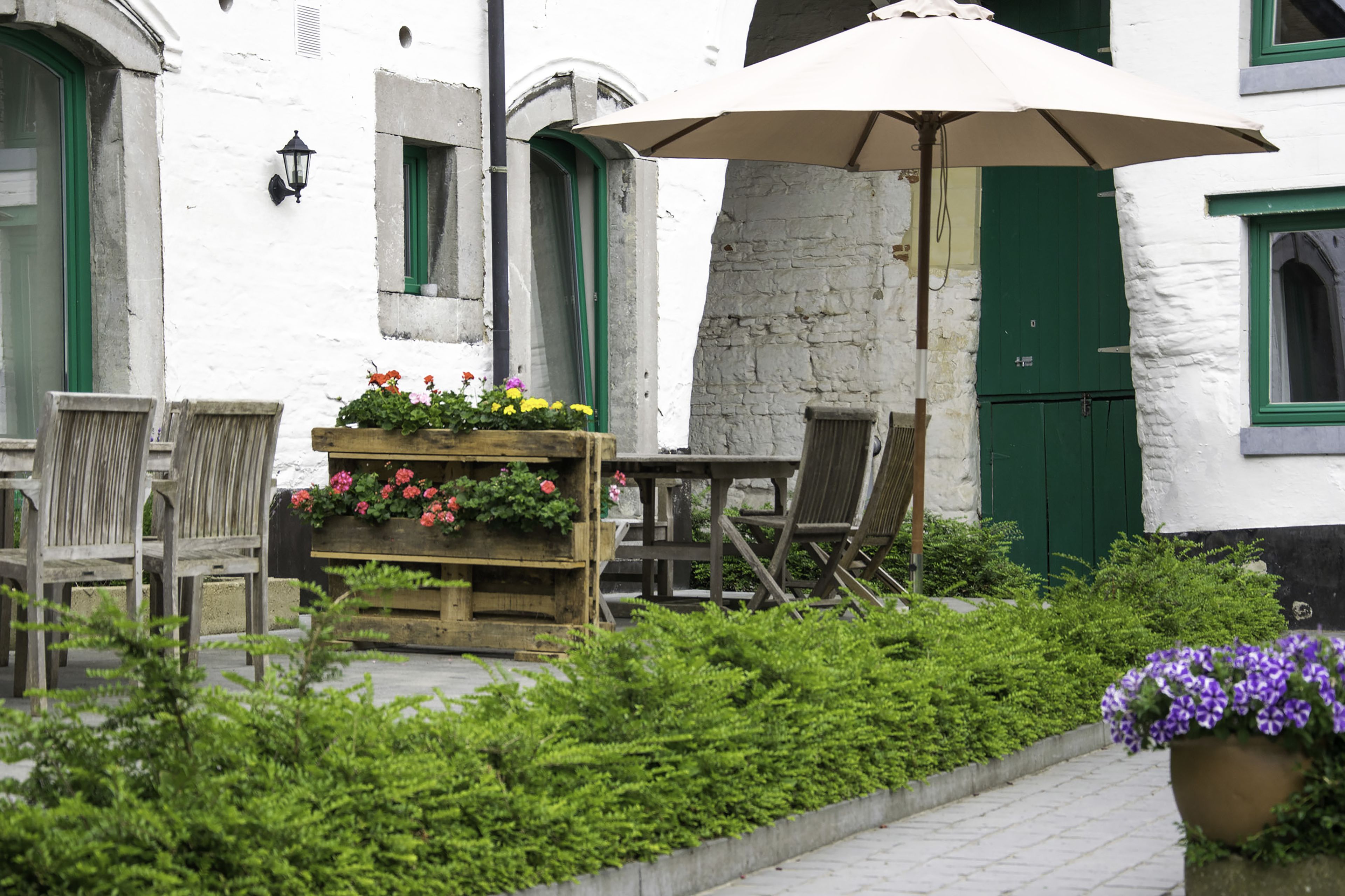Een terras met zitplaatsen buiten, een plantenbak met bloemen, en een gebouw met groene luiken.