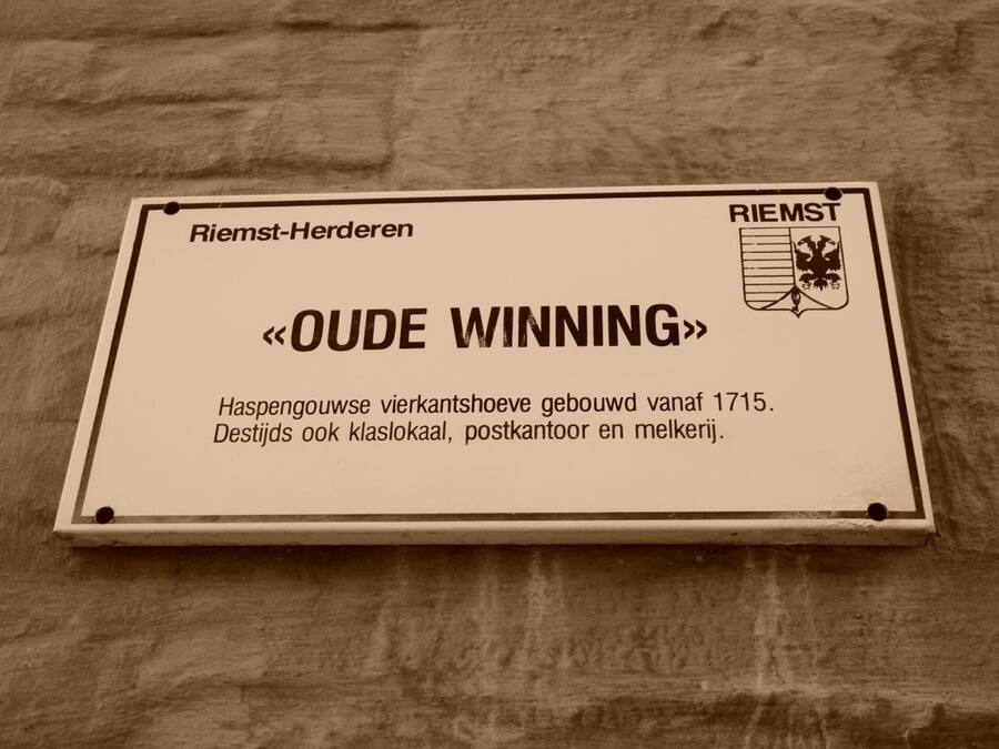 Een bord met de tekst 'Riemst-Herden' en 'OUDE WINNING' op een muur.