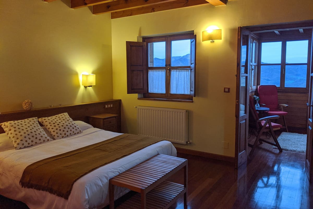 A bedroom with a bed, bedside tables, a bench, a window with curtains, and a view of the outdoors.