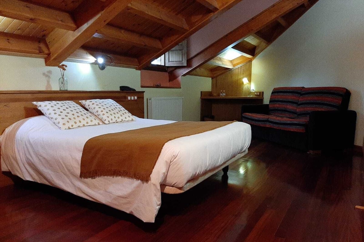 A bedroom with a wooden ceiling, a bed with a polka dot pillow, a striped chair, and a small table with a lamp.