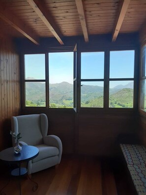 A room with a wooden ceiling, a sofa, a small table with a vase, and a bed with a patterned cover. Large windows offer a view of green hills and mountains. - El Rexacu (Onis)