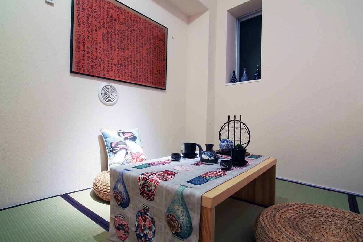 A traditional Japanese room with tatami flooring, a low table with a tablecloth, a woven chair, and a window with a view.