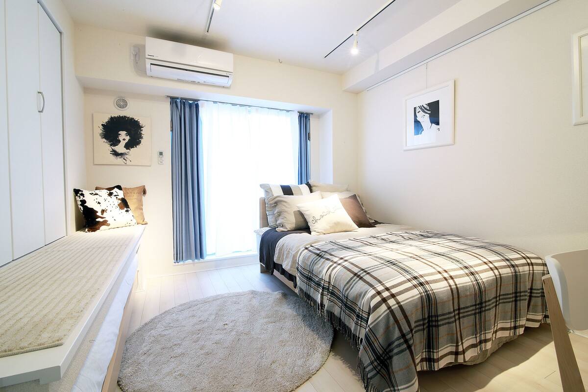 A bedroom with a bed, a bedside table, a window with curtains, a wall-mounted air conditioner, and a wall art piece.