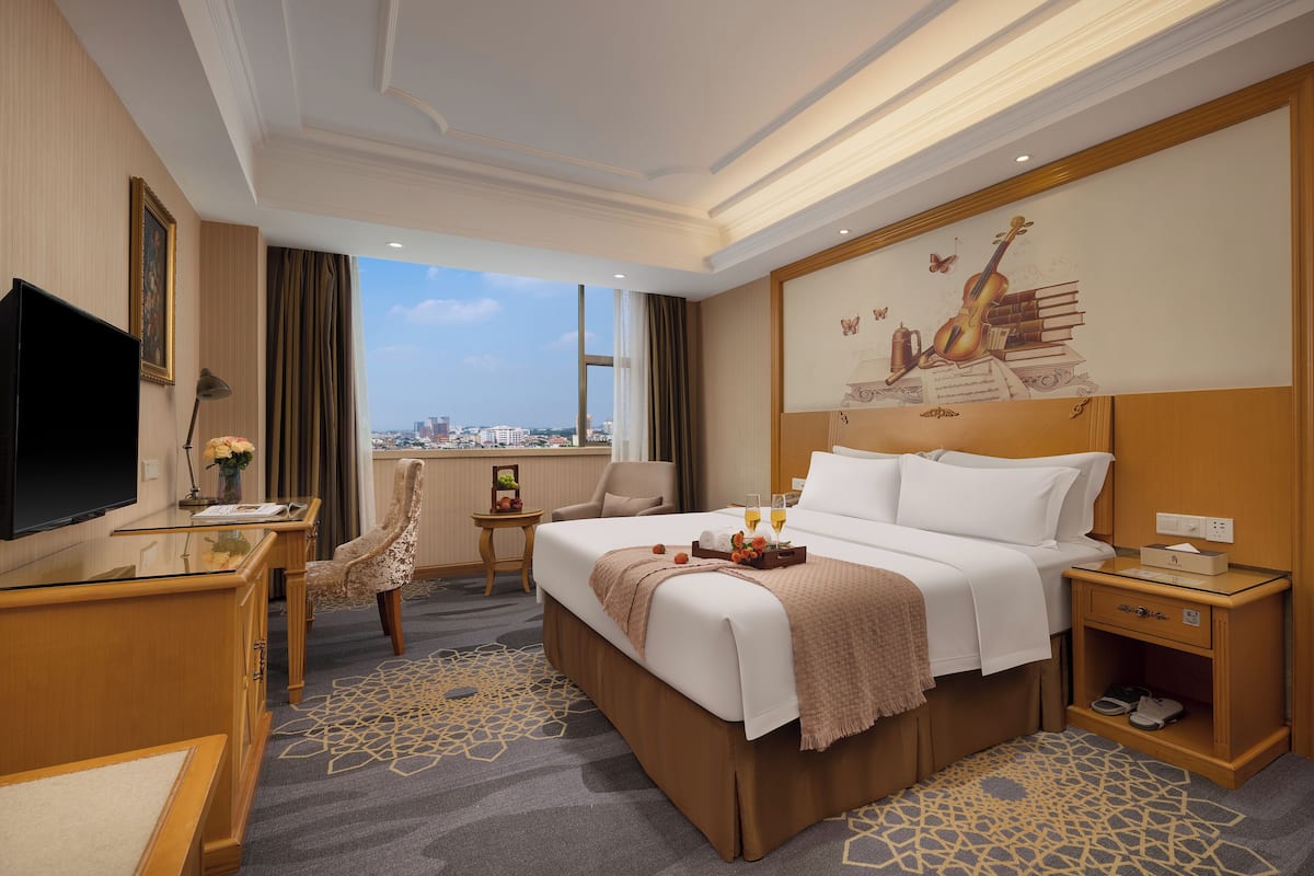 Habitación de hotel con una cama grande, un escritorio con televisor de pantalla plana, una silla, una mesita con un jarrón de flores y vistas a la ciudad a través de amplias ventanas.