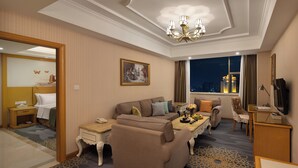 Habitación de hotel con sofá, sillón y una mesita con lámpara. Se observa una cama con mesita de noche, un espejo y una ventana con vistas a la ciudad de noche.