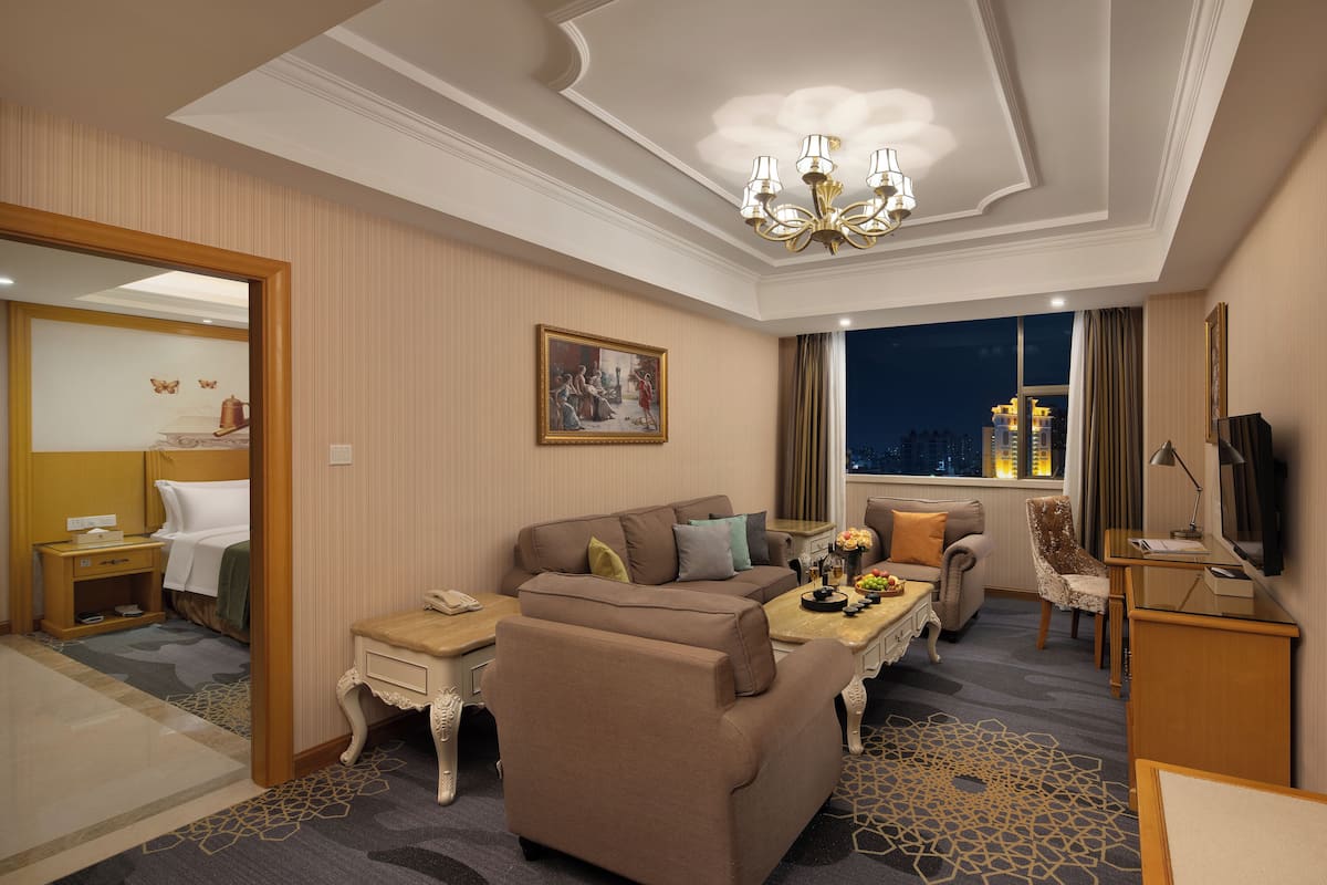 Habitación de hotel con sofá, sillón y una mesita con lámpara. Se observa una cama con mesita de noche, un espejo y una ventana con vistas a la ciudad de noche.